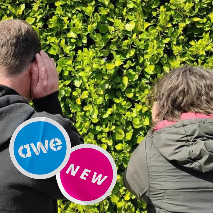 A man and a woman face away from the camera, towards a hedge with bright green leaves. They are cupping her ears, as if to listen closely. Graphics of stickers have been overlaid on the bottom-left corner of the image, one showing the acronym ‘AWE’ (for Art Week Exeter), the other showing the word ’new’. (Image credit: Gem Smith, Take-A-Part.)