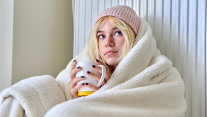 Blonde girl wearing a pink beanie. She is wrapped in a blanket holding a mug. 