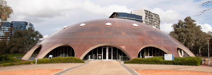 The Shine Dome in Canberra is shown with wide archways beneath a shiny copper rooftop with evenly spaced circular white skylights. Three taller buildings are in the background.