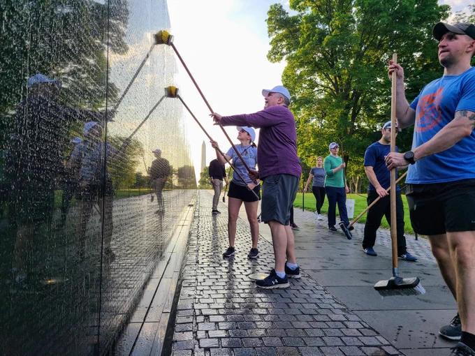 A small group of men and women in work out cloths and hats use long handled brushes to clean a long black stone memorial wall.
