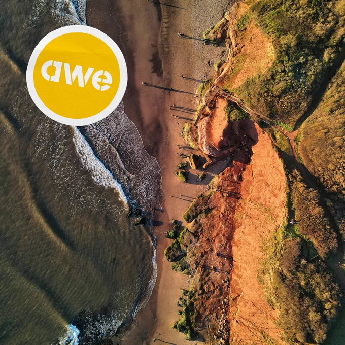 An aerial photo the beach at Orcombe Point, Exmouth, Devon, UK. The sea gently laps the beach from the left, to the right there are red cliffs with green vegetation on top. A small number of walkers appear as tiny dots in the picture, casting long shadows in the low sunlight. The Art Week Exeter ‘awe’ logo appears on the graphic of a sticker overlaid on the image. (Image credit: #RedZeppelin via @unsplash.)