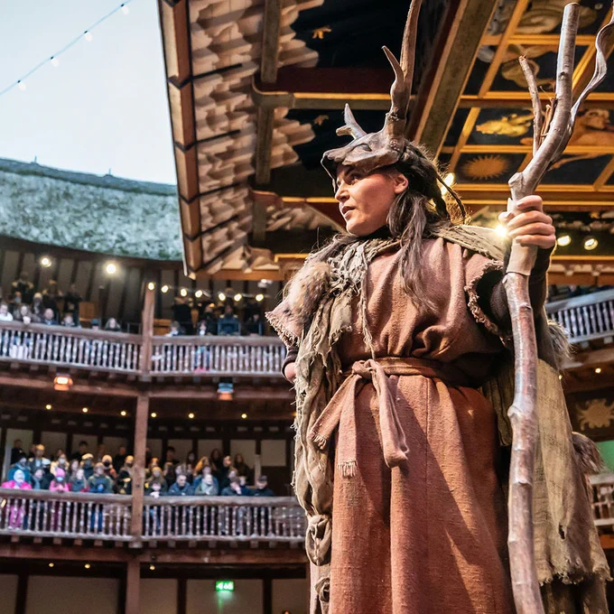 An actor wearing brown rags and a fur cloak, with an animal antler headpiece, holds a wooden staff on the Globe Theatre stage, looking up at the audience sat in the circular Galleries around them.
