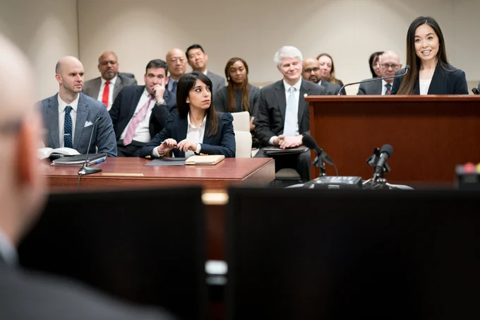 A woman speaks at a podium in a courtroom while a group of people look on