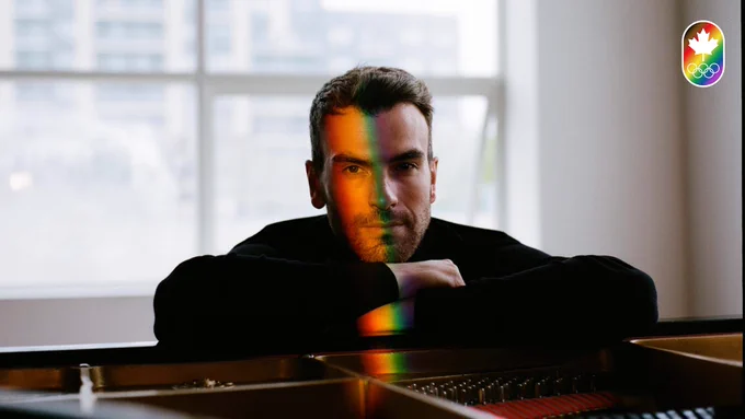 Eric Radford leans on a piano with a rainbow light shining down his face