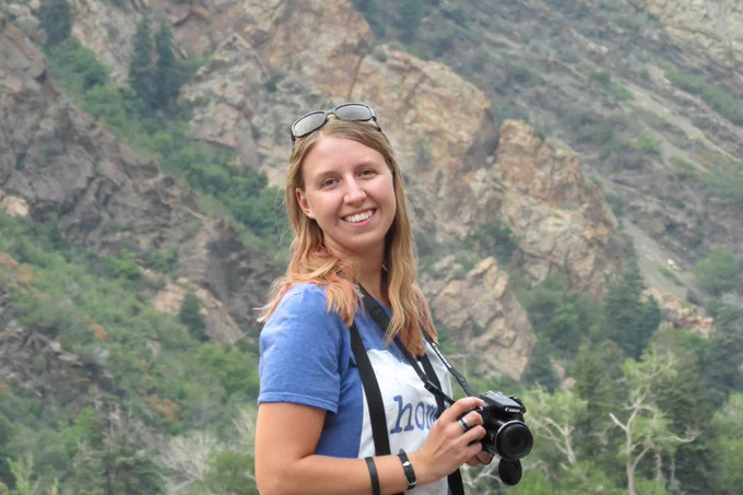 Sarah, a blond long-haired person, wearing a blue and white shirt and black sunglasses and carrying a camera, in front of some mountains in Utah