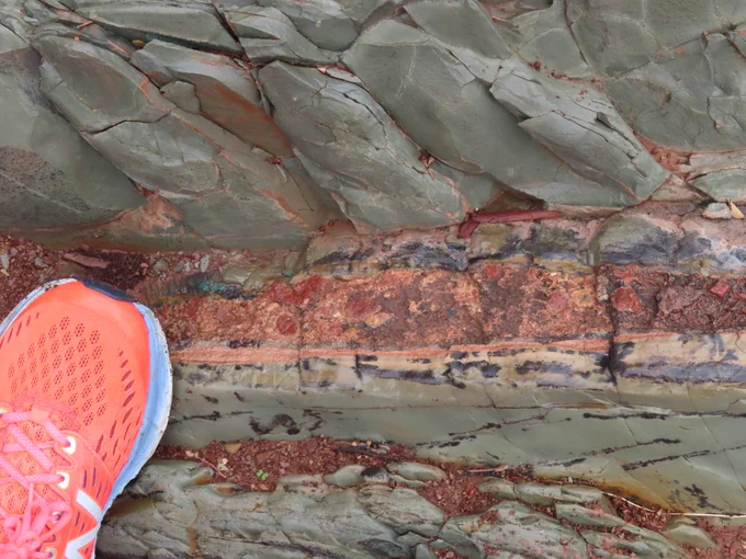 Photo of a rock, with a neon orange shoe for scale. The top and bottom of photo are green-grey, there is a distinct layer across the middle of orange colour rock, about 3-5cm thick. It contains some cm-scale red colour clasts. A patch of green copper mineralisation can be seen above the ejecta layer.