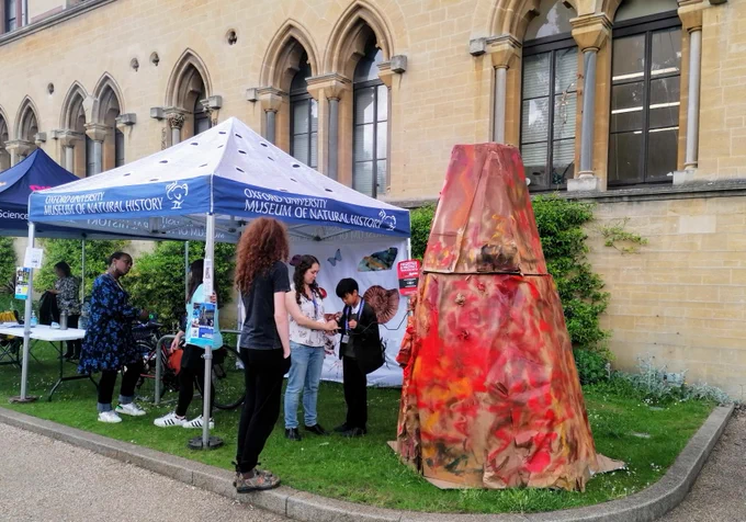 A group of people look at a 'volcano' constructed from painted cardboard (and with a smoke machine inside!) on the front lawn of the Oxford University Museum of Natural History. Some people stand under a gazebo.