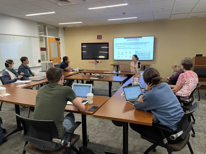 Allison Xu talks to a group of faculty and graduate students about data in a modern and well light classroom. Behind her is a screen with a slide that explains unstructured data.