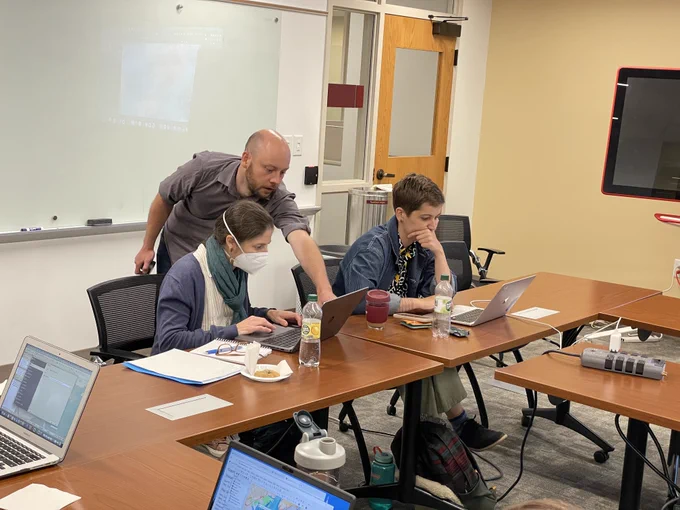 Matt Naglak stands behind two faculty members who are working intently on laptops. He's leaning forward and pointing at one of the laptop screens as he instructs one of the faculty members on how to use QGIS, a mapping tool. 