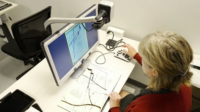 Our SET-BC educator, Naomi, sits at a desk with the DaVinci video magnifier, in front of her. The magnifier's long arm extends over her page on the desk.  The image of the page is on the screen in front of her.