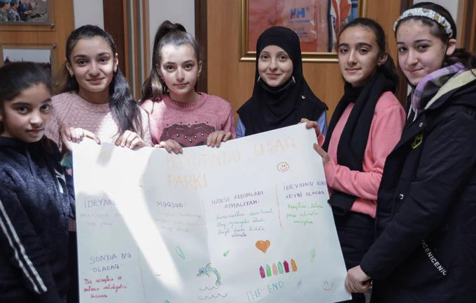 Five adolescent girls are standing in a room, holding a poster they designed. They are all smiling and looking directly into the camera.