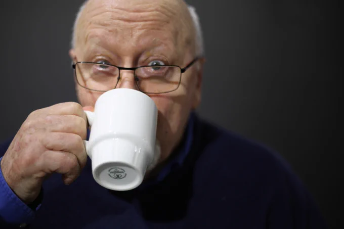 Image of an older man looking to camera sipping from a white cup