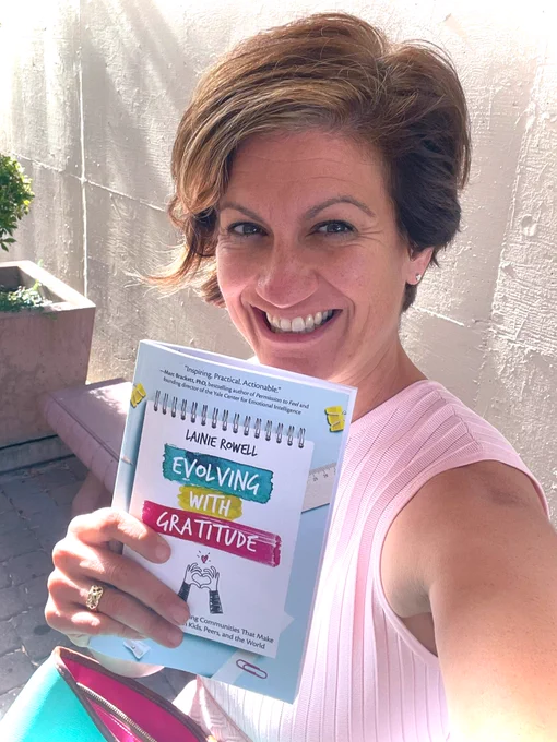 Katie Novak, wearing a pink dress, holding the book Evolving with Gratitude by Lainie Rowell.