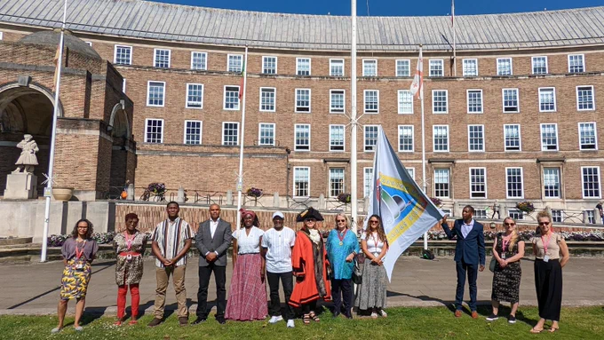 City Hall is in the background, with a crowd of people stood on the grass of College Green. Forward Maisokwadzo holds the Windrush flag, before it is raised.