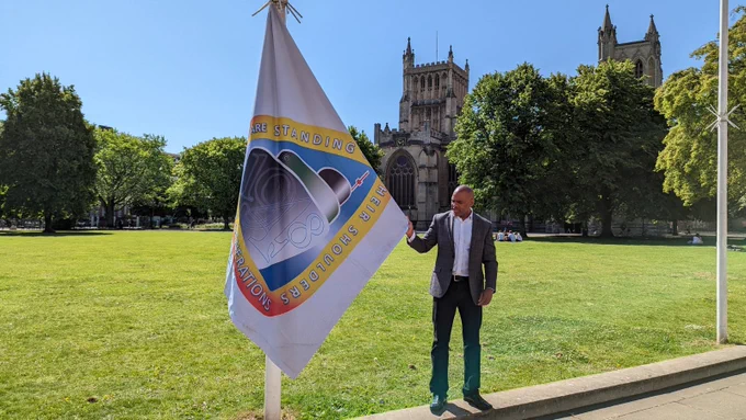 Mayor of Bristol Marvin Rees stands on College Green in the sun in front of Bristol Cathedral. He is standing next to a flagpole and is holding the Windrush flag. The flag is a ship on a blue background surrounded by white text on an orange border. The ship says '1948'.