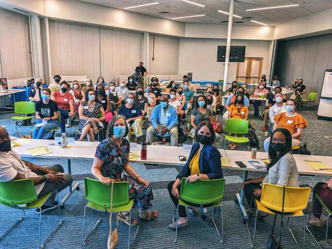 An image of 46 majority camera-facing, medically-masked educators and community members standing and seated throughout a conference room with the 5 closest to the camera seated at a long rectangular table before the rest watching as a standing and seated audience at EdCo Collective Conference. 