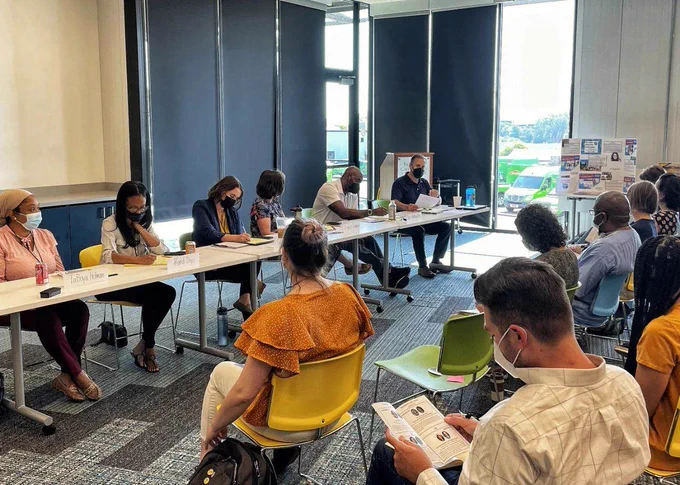 An image of 6 seated educators and panelists presenting from a long rectangular table with 8 seated educators and audience members watching them, the majority of all wearing medical masks, in a windowed conference room at EdCo Collective Conference. 