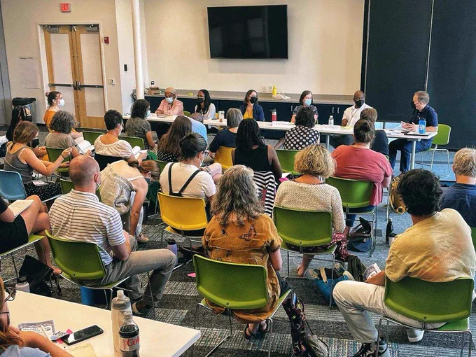 An image of 6 seated educators and panelists beneath a large wall-mounted screen presenting from a long rectangular table before 22 seated educators and audience members, the majority of all wearing medical masks, in a conference room at EdCo Collective Conference.