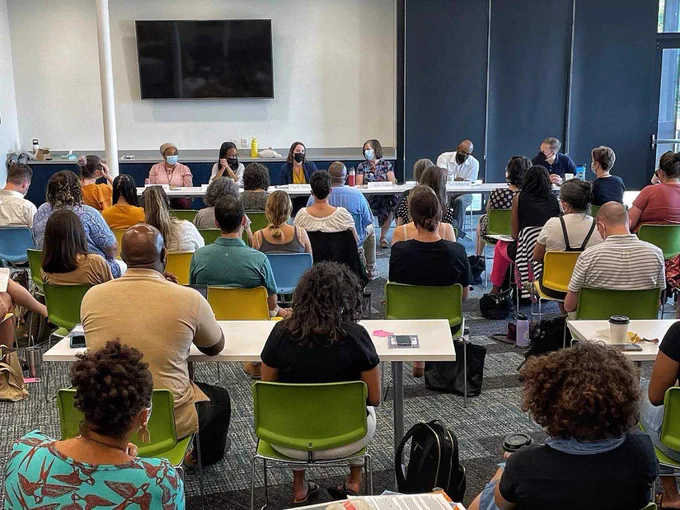 An image of 6 seated educators and panelists, all wearing medical masks, beneath a large wall-mounted screen presenting from a long rectangular table before 28 seated audience members, 3 of whom are seated at small rectangular tables, in a windowed conference room at EdCo Collective Conference.