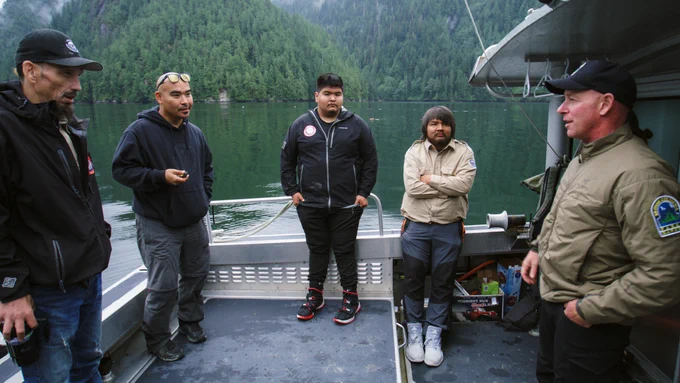 Elected Kitasoo Xai'xais chief Doug Neasloss stands on an aluminum boat with his team of uniformed coastal guardian watchmen and a BC Park Ranger having a friendly discussion on a calm day with the green waters of a high-cliffed inlet in the background.