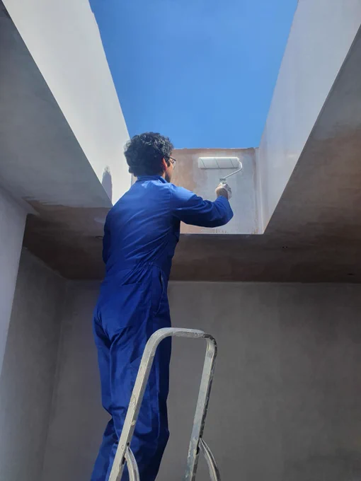 A volunteer stands on top of a ladder painting a skylight, with the bright blue sky visible through it