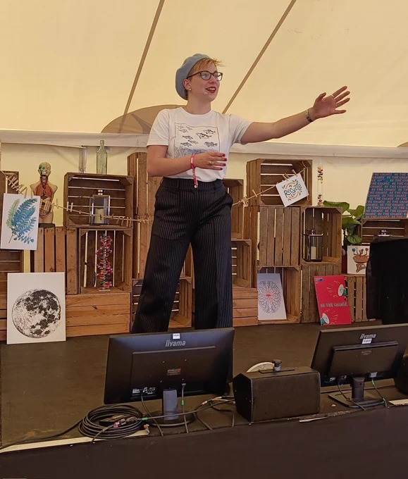 Me on a stage, wearing blue suit trousers and a white shirt with blue fish printed on it. I'm also wearing a small mic and a blue beret. My arms are gesturing to something off-screen.