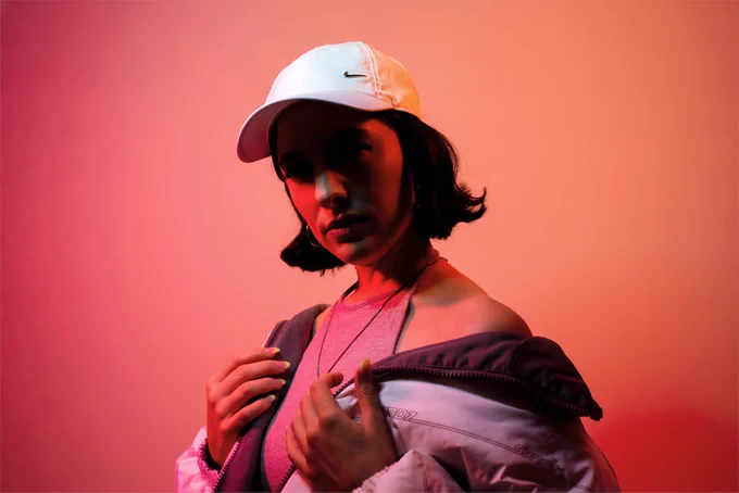 FOURA poses with her arms resting on her jacket. She is bathed in red light and her face is partially obstructed by the shadow cast by her white cap.