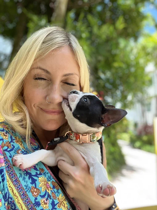 Kelly with eyes closed smoking, holding Freya the Boston terrier puppy. Freya is biting Kelly’s cheek