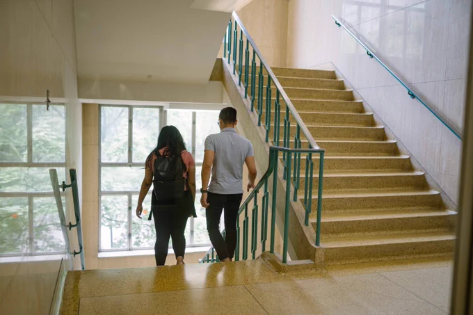 Two students walk down a staircase towards large glasses windows in Toronto Metropolitan University's Kerr Hall.