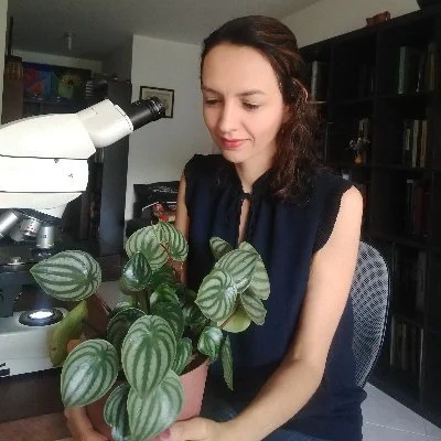 A picture of Natalia wearing a dark blue shirt, sitting next to a microscope and looking at a green plant