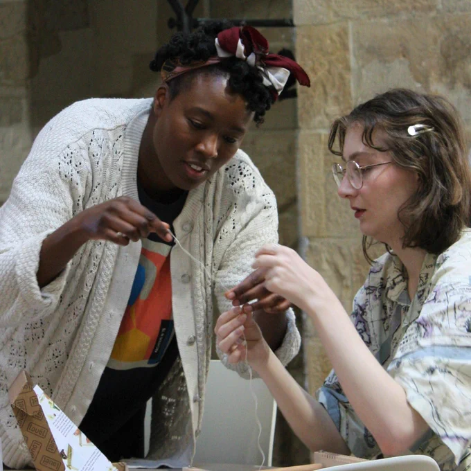 Woman standing helps woman sitting at table tie a knot using a strand of yarn. 