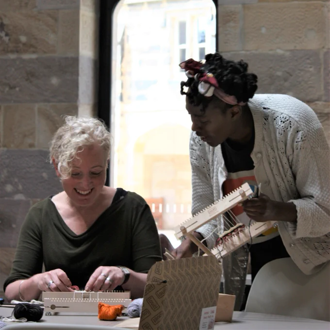 Woman standing shows woman sitting her small hand-held loom. 