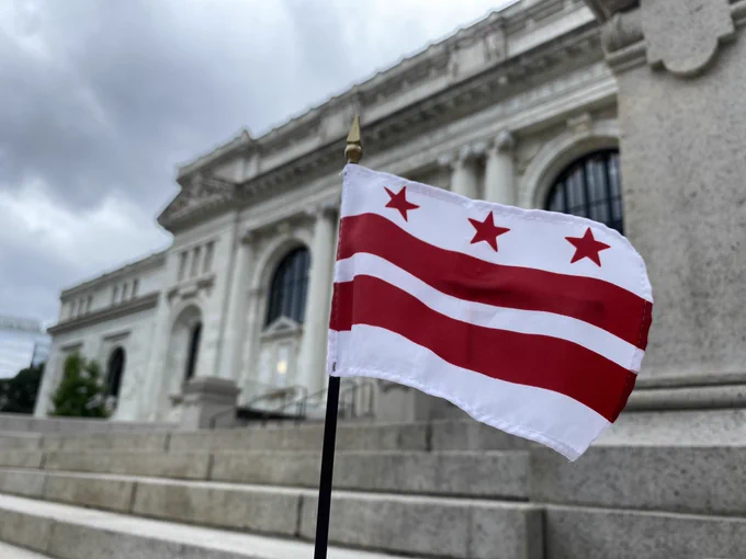 The flag of Washington, DC consists of 3 red stars above 2 red bars on a white background. In the background is Carnegie Library, home to the DC History Center in Mount Vernon Square.