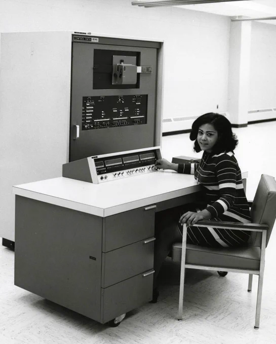 CBI Image of the Day. A woman working at an 8090 Control Data Corporation CDC 8090 in the late 1960s, likely 1967 or 1968. It seems oddly isolated in a fairly empty large room. She is looking at the camera turning sideways with her right hand on the console that sits on a white top and steel drawer desk/computer station on wheels. Behind this is main console of the 8090. There is a white tile floor. The control panel is tilted and has a black face with silver edges.