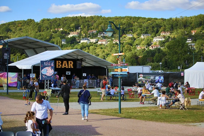 Au premier plan, des visiteurs se promènent dans le village d'un festival musical. Arrière plan : des maisons dans la verdure yvelinoise.