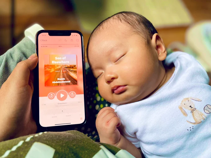 A baby sleeps beside on a breastfeeding pillow. Beside his head, his mother holds a phone displaying the audiobook “Son of Elsewhere” by Elamin Abdelmahmoud.
