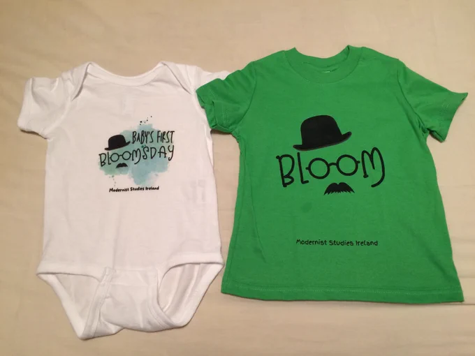 Photo shows a short-sleeved white baby bodysuit reading 'BABY'S FIRST BLOOMSDAY' and a green children's t-shirt reading 'Bloom'. Both items also say 'Modernist Studies Ireland' at the bottom, and the o's of the 'Blooms' are like glasses with a Joycean moustache below and hat above.