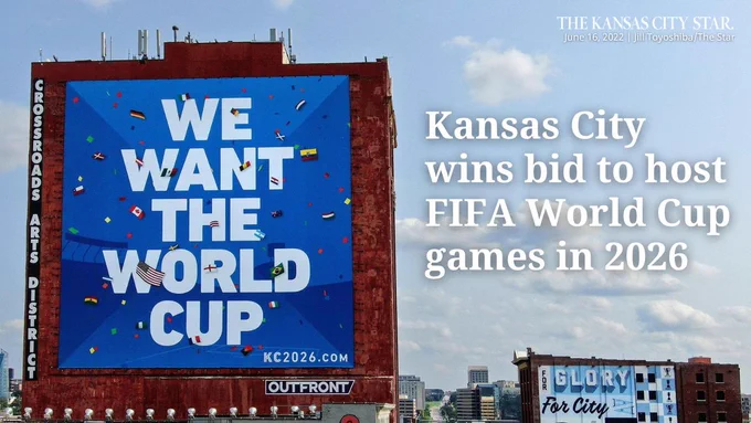 A 90 foot by 90 foot banner hangs off the side of a Kansas City building that reads "We want the World Cup." Text on the card reads, "Kansas City wins bid to host FIFA World Cup games in 2026"