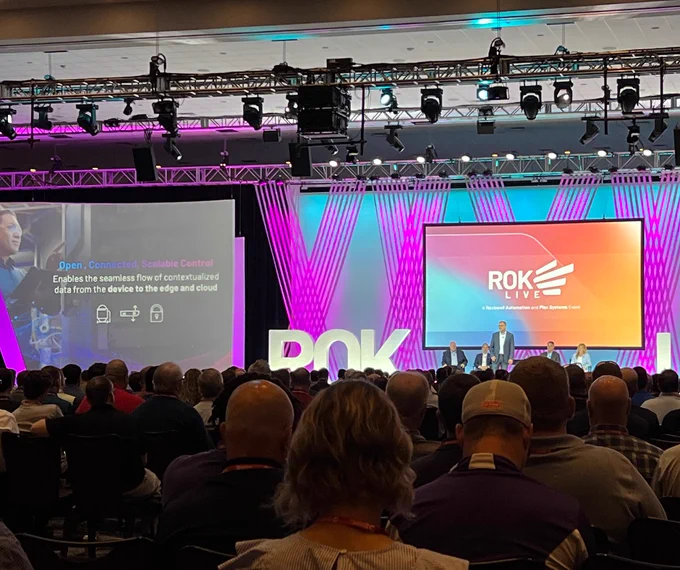 A speaker on stage in front of an audience. There is a big projector screen with the ROKLive logo.