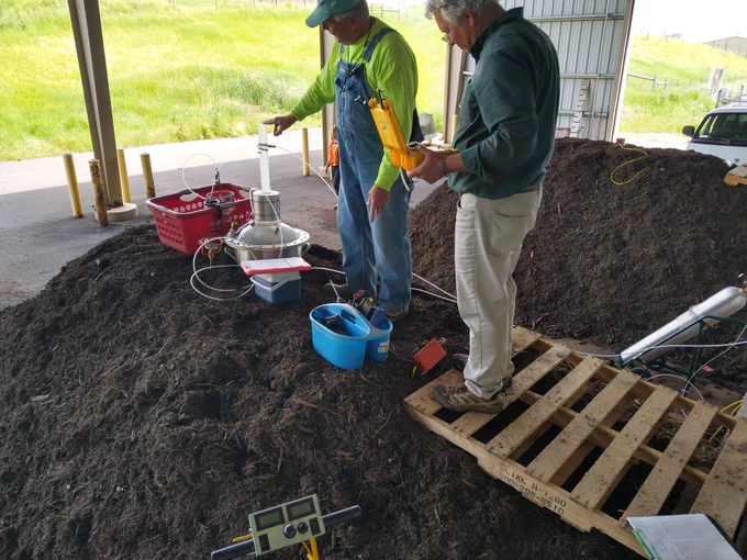 Prof Jobson and Dr. Schmidt stand on a compost pile and conduct real-time measurements of temperature, ammonia, carbon dioxide and helium (used as a tracer) in the flux chamber stack while a canister and impinger collect an integrated whole air sample for later analysis.