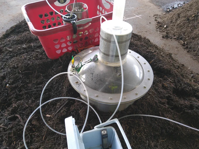 View of a flux sampling dynamic chamber with attached collection chamber and impinger. Condensation from the hot compost is visible on the chamber surfaces. A tube delivers clean air with a helium tracer into the chamber. 