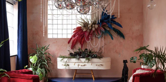 White coffin surrounded by colourful feathers, disco balls and plants. 