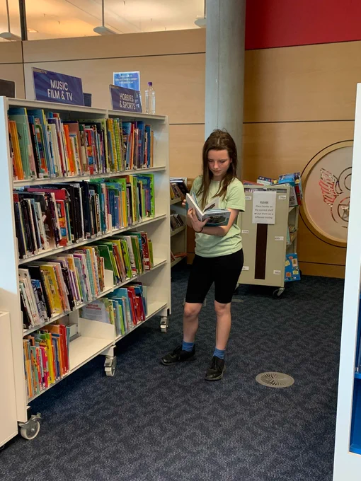 A Year 7 student stands looking down at the open book she is holding. Beside her is a shelf on wheels topped by signs reading "Music, Film & TV".