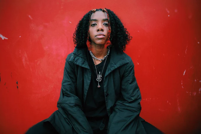 Dua Saleh is wearing all black. They have on a black athletic kimono button up coat with a hoodie and all black clothes. They have their natural hair out. They have two red and black braids in front and curly hair in back . They have on silver jewelry: a link chain, a Scorpio chain, and a dragon claw with a stone in it. Their crouching with a red background behind them. ❤️