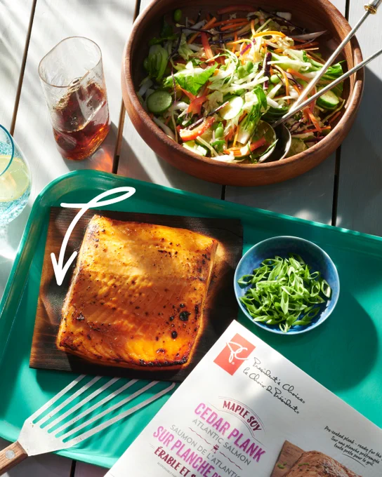 A piece of salmon on a wood plank served onto a tray. The table is also set with a fresh summer salad, a half-full glass of iced tea and a small bowl of sliced green onion. The box for PC Maple Soy Cedar Plank salmon is in bottom corner. 