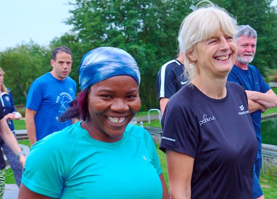 Two women wearing activewear are smiling while walking in a park. There are people in the background behind them.