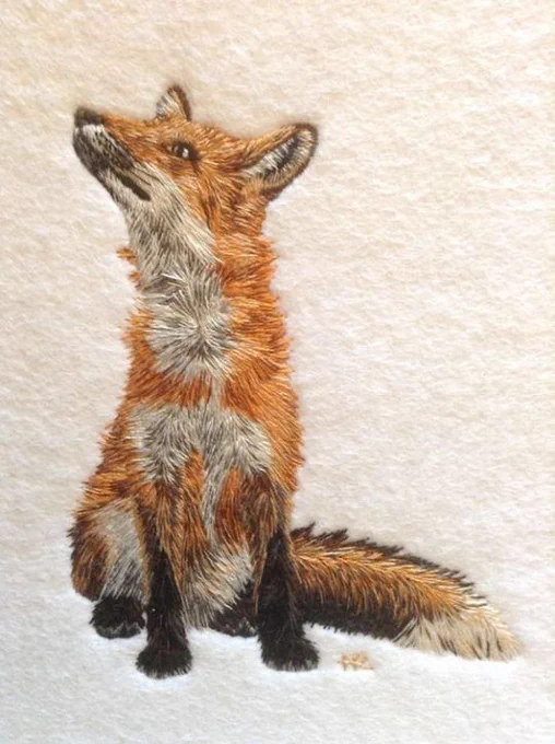 Embroidery in a pictorial style featuring a ginger, white and brown sitting fox, facing forwards and head turned up to the right on a plain cream background 