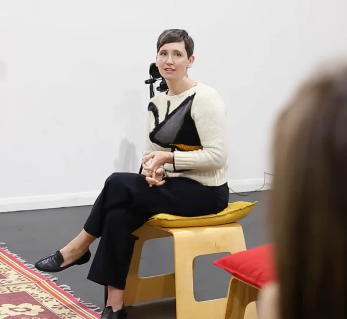 Image of Artistic Director Dr Jessica Olivieri sitting on a short brown stool in a large white room in front of a colourful rug.
