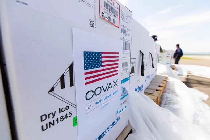 Boxes of vaccines with the COVAX logo and U.S. flag sit on a tarmac in Mongolia as two airport employees offload them from palettes in the distance.
