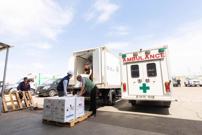Photograph of four men loading boxes of COVID-19 vaccines with the COVAX logo and U.S. flag onto a white truck. A white ambulance with a green cross is parked next to the larger white loading truck.